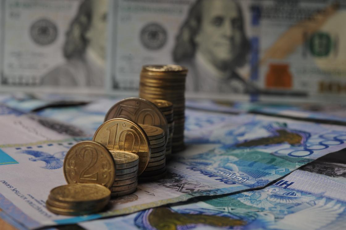 Стопки монет тенге стоят на фоне купюры доллара