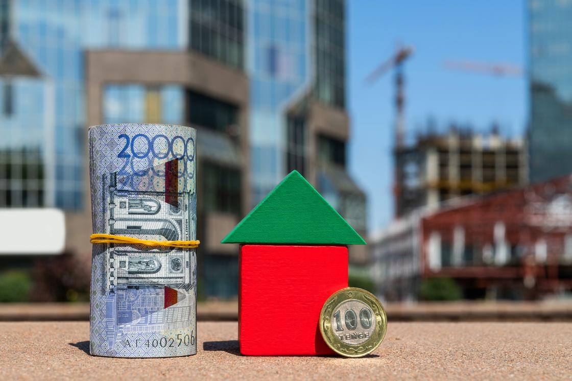 Свернутая купюра 20 тысяч тенге и монета 100 тенге стоят рядом с кубиками в виде дома Свернутая купюра 20 тысяч тенге и монета 100 тенге стоят рядом с кубиками в виде дома