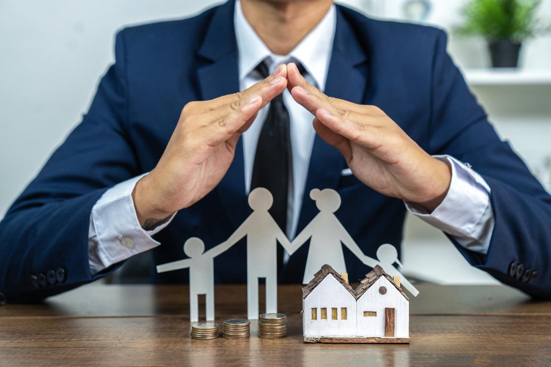 Мужчина закрывает руками человеческие фигурки и игрушечный домик Мужчина закрывает руками человеческие фигурки и игрушечный домик